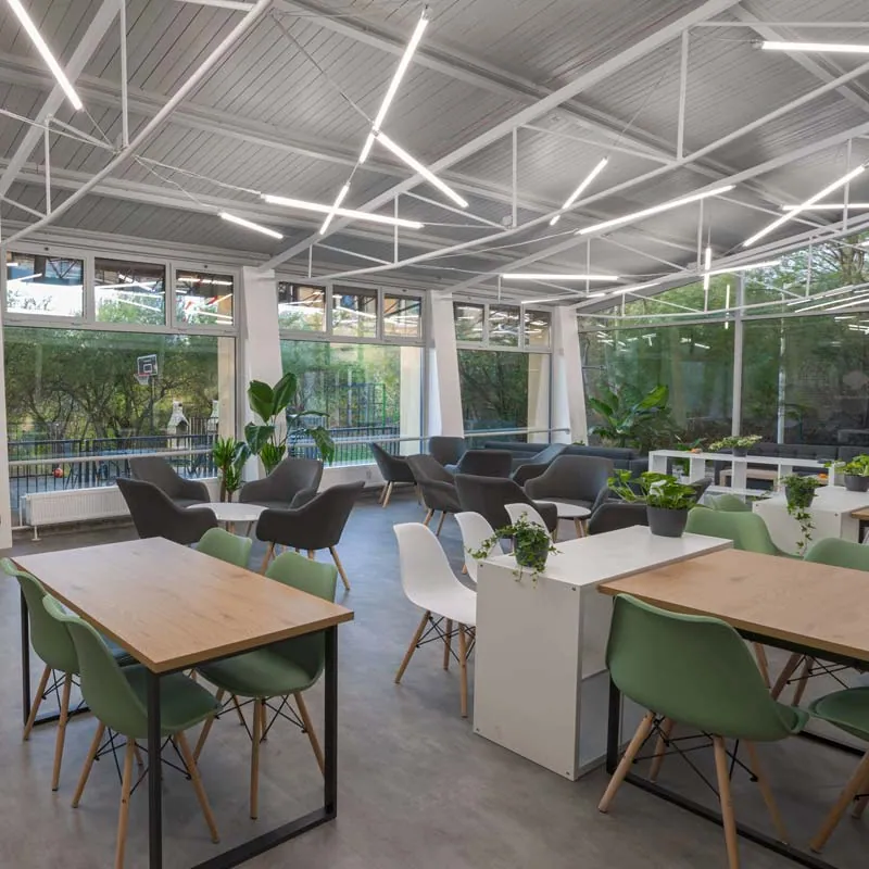 Bright glass-walled Bro-coli lounge with wooden tables, green chairs, armchairs and plants