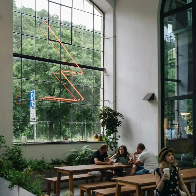 People at wooden tables in a café with tall industrial windows and a neon sign